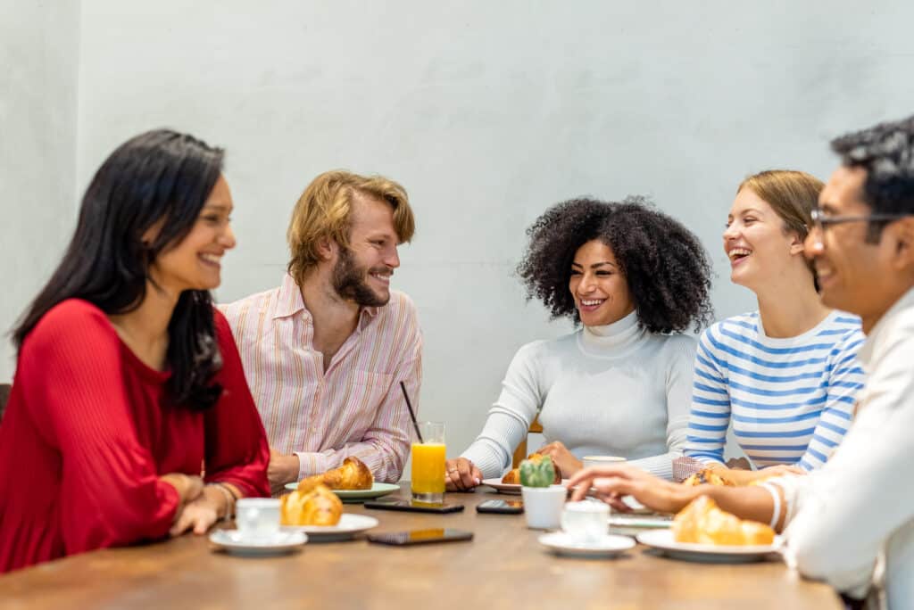 Group of multi ethnic professionals having brunch, focus on the blonde man and the afro hair woman, table with coffee, cappuccino, orange juice and pastry.